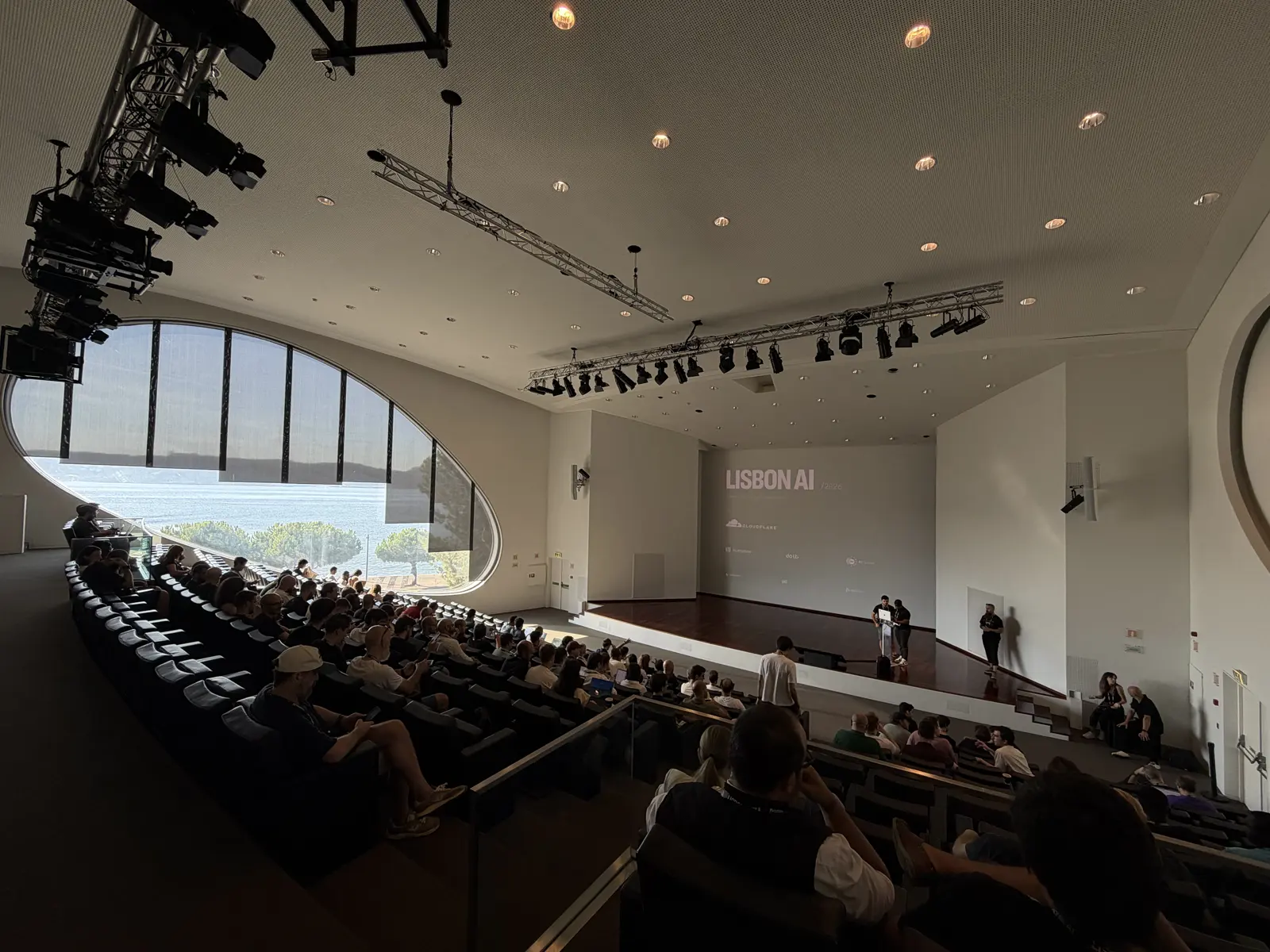 Auditório em anfiteatro quase cheio, com uma grande janela oval à esquerda e o palco ao fundo.