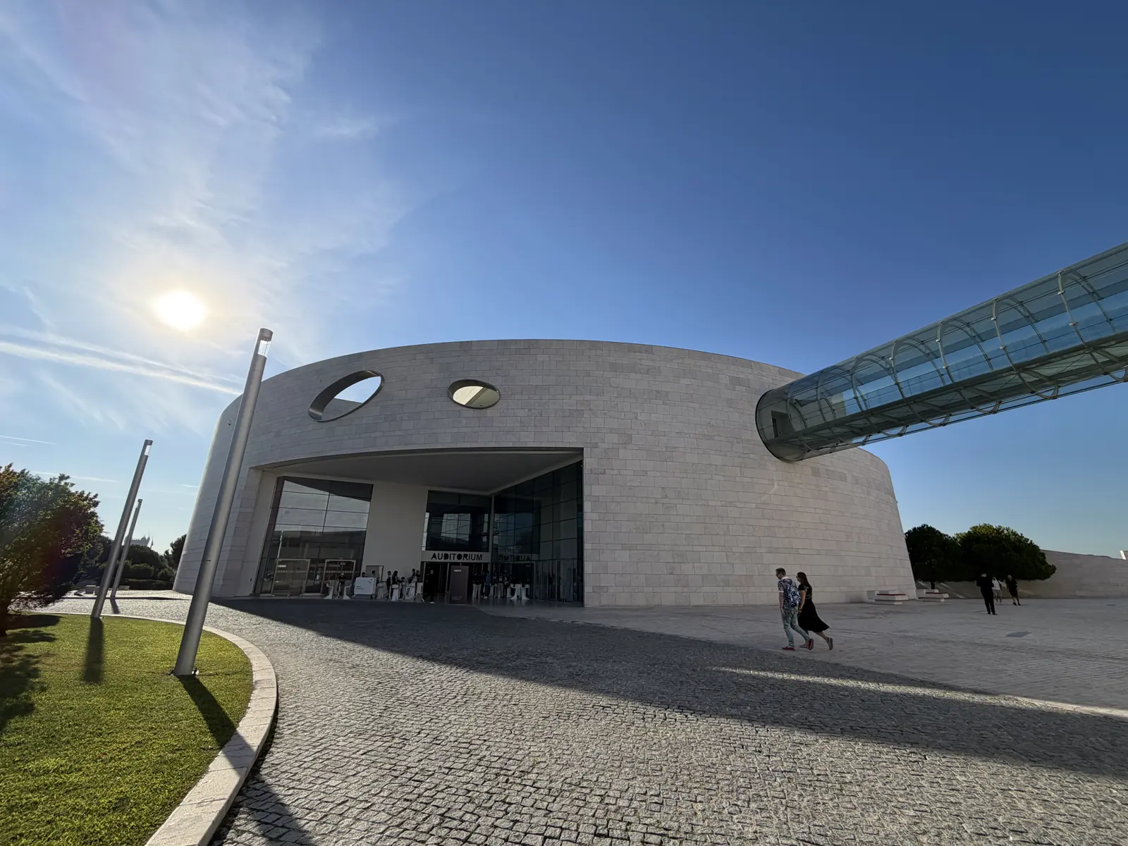 Fachada curva do Centro Champalimaud sob céu azul, com uma passarela de vidro saindo do prédio e o sol no alto.
