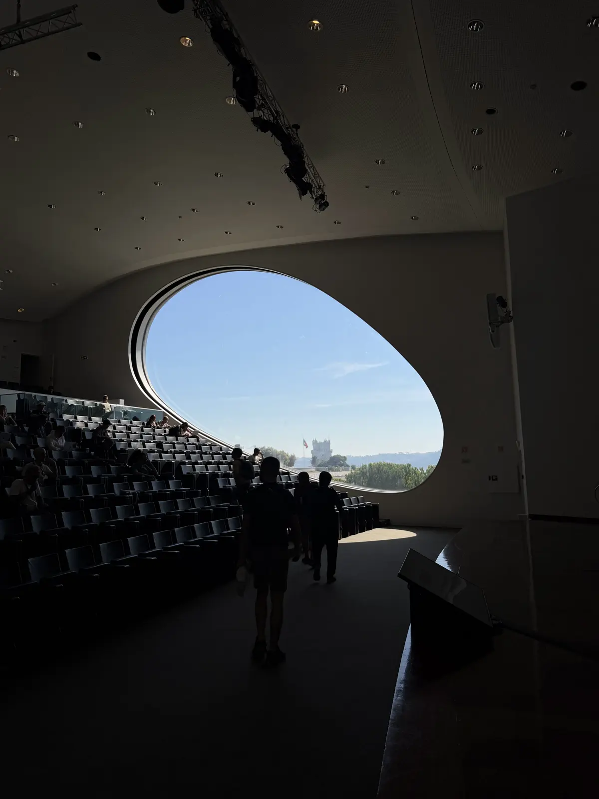 Auditório escuro e vazio, com a luz entrando pela janela oval e a silhueta de uma pessoa descendo a escada.