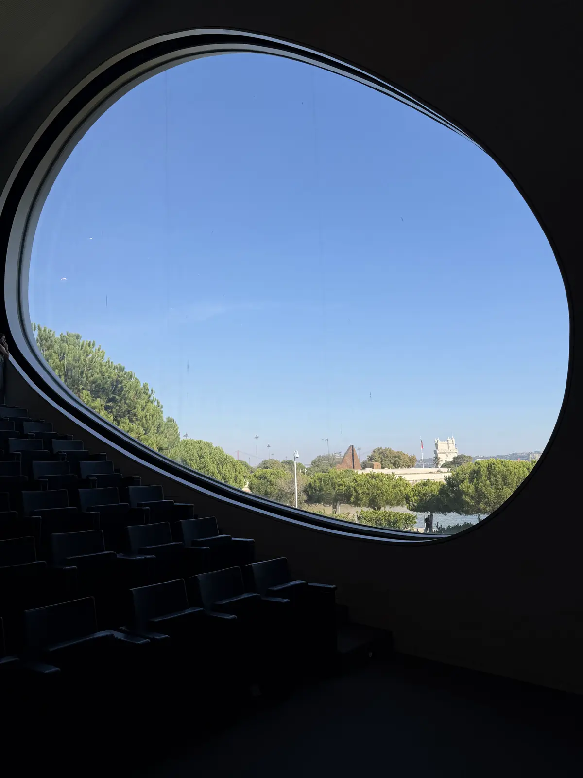 A janela oval vista de dentro do auditório escuro, emoldurando árvores e céu azul.