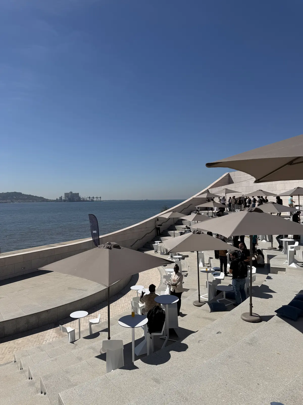 Terraço com guarda-sóis e mesas cheias de gente, com o rio ao fundo.